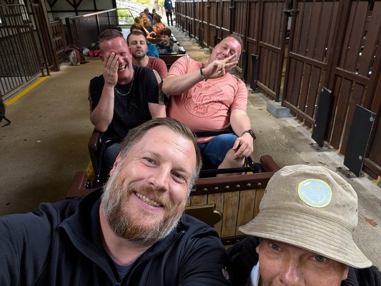 Eine Gruppe von Männern sitzt in einem Wagen einer Holzachterbahn. Im Vordergrund machen sich zwei Männer ein Selfie und lächeln breit. Dahinter reagieren zwei weitere Fahrgäste mit Lachen und Gesten vor Aufregung auf die Fahrt.