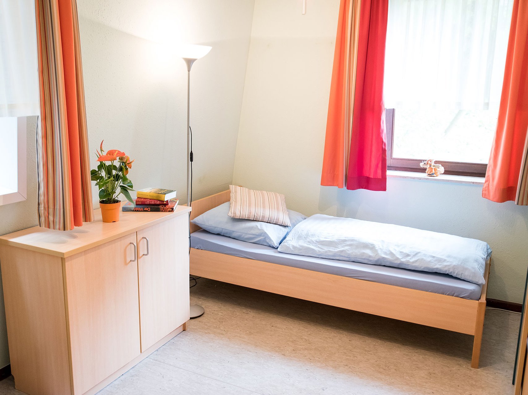 Bewohnerzimmer mit Bett und Schrank - hauptsächlich in orange gehalten und sehr hell