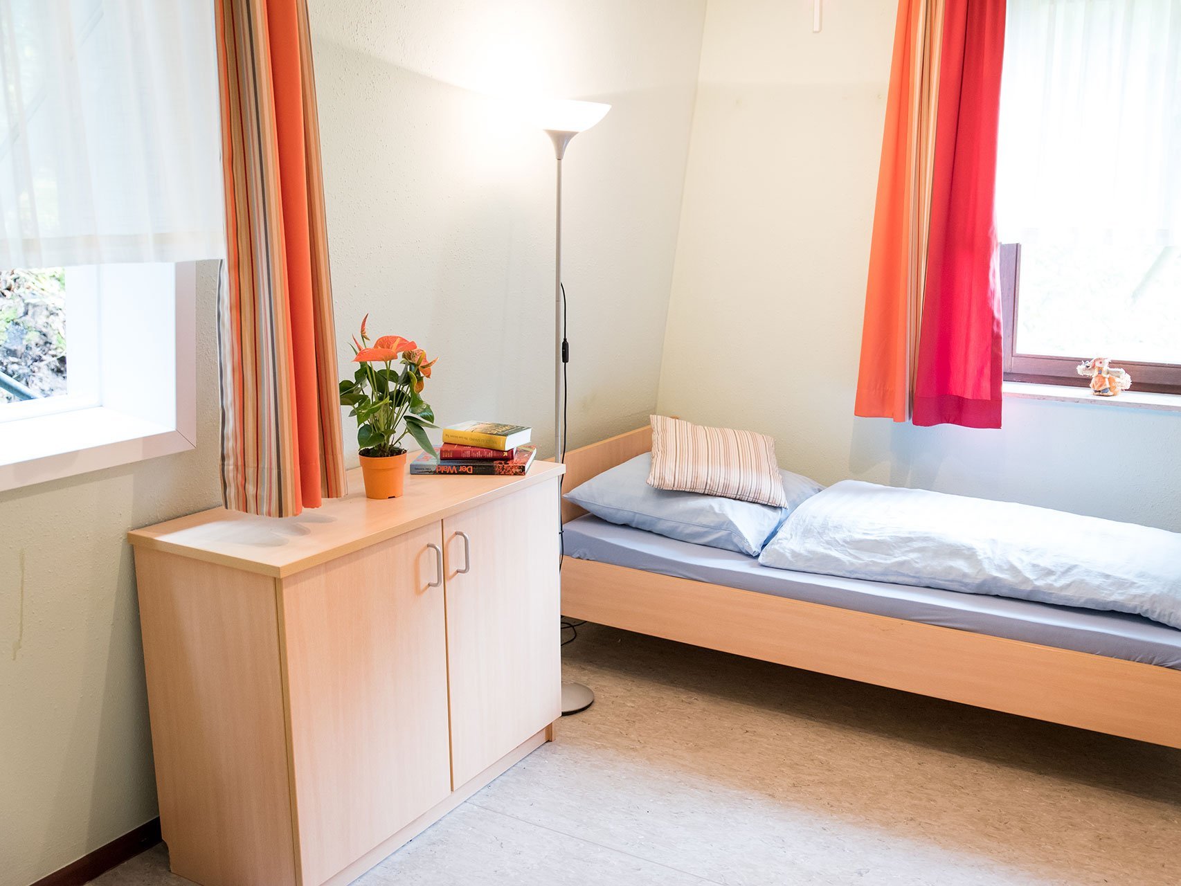 Bewohnerzimmer mit Bett und Schrank - hauptsächlich in orange gehalten und sehr hell