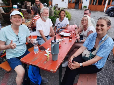 Eine fröhliche Gruppe sitzt an einem langen roten Tisch im Freien. Eine Person links zeigt einen Daumen hoch, während eine Frau rechts lächelt. Auf dem Tisch stehen Getränke und eine rosa Schweinestatue. Die Szene wirkt entspannt und gesellig.